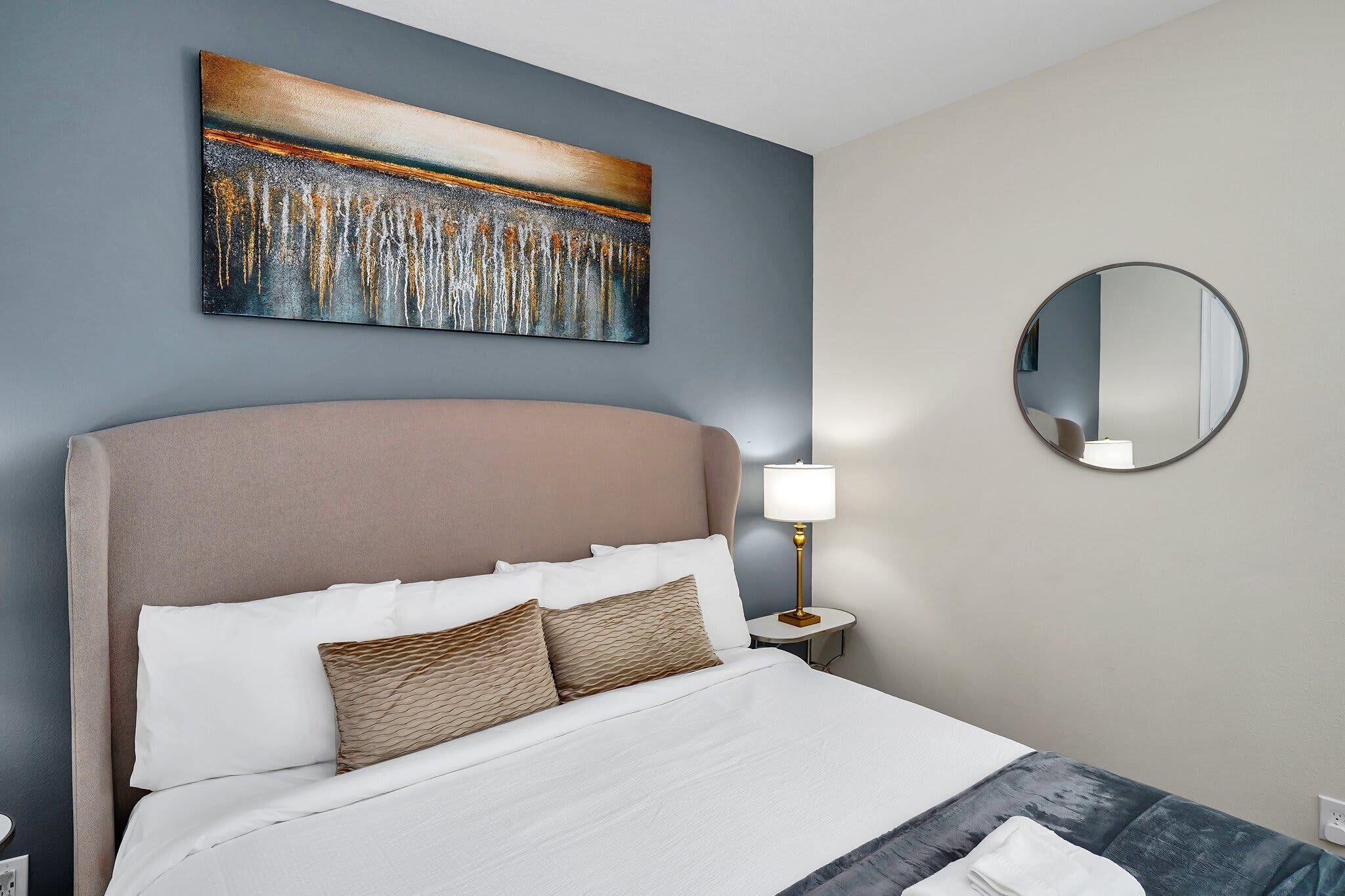 A neatly made bed with white linens and two patterned pillows, a bedside lamp, and a large abstract painting on the wall.