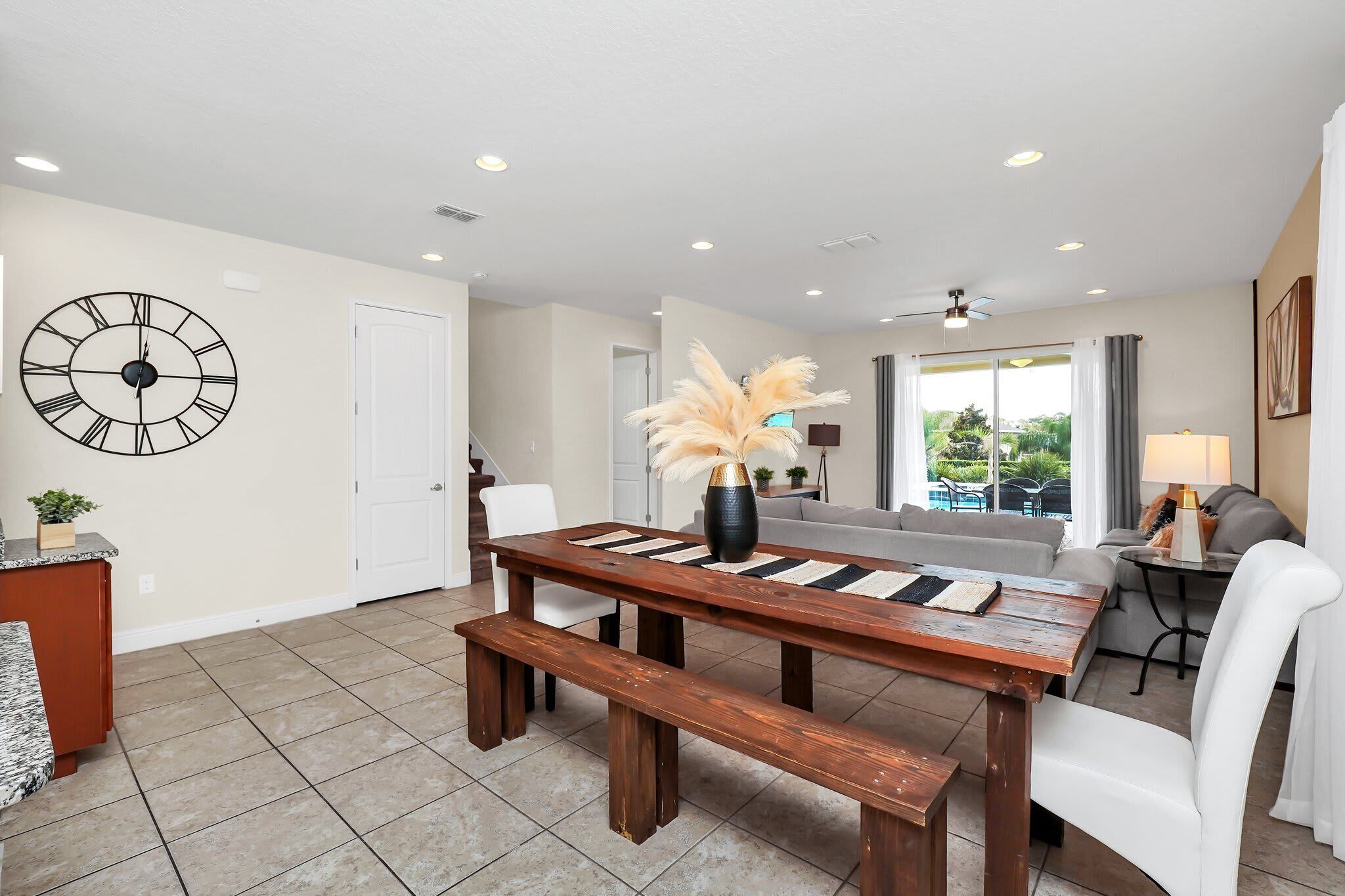 A spacious living area with a wooden dining table, benches, a large wall clock, and a view of the outdoors.