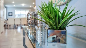 Une plante en pot, placée sur une surface réfléchissante dans un espace intérieur moderne, agrémentée d’un élément décoratif mural et laissant entrevoir un coin repas à l’arrière-plan.