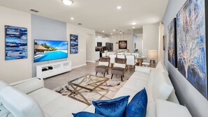 Un salon moderne avec un canapé blanc, une table basse et un téléviseur. On aperçoit à l’arrière-plan un coin repas avec des chaises et une table.