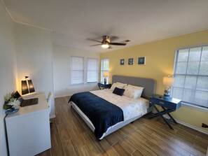 A bedroom with a bed, bedside tables, a desk, a ceiling fan, and windows with blinds.