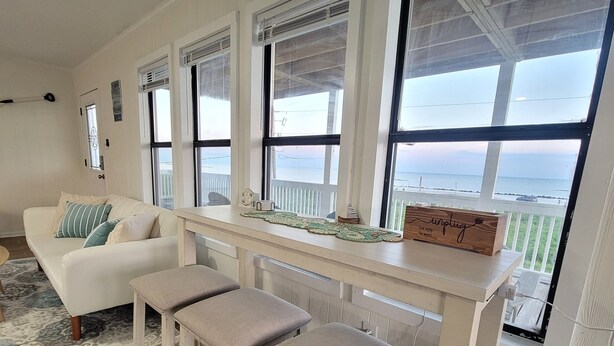 A coastal living room with a white sofa, two cushioned stools, a wooden table with decorative items, and a view of the ocean through large windows.