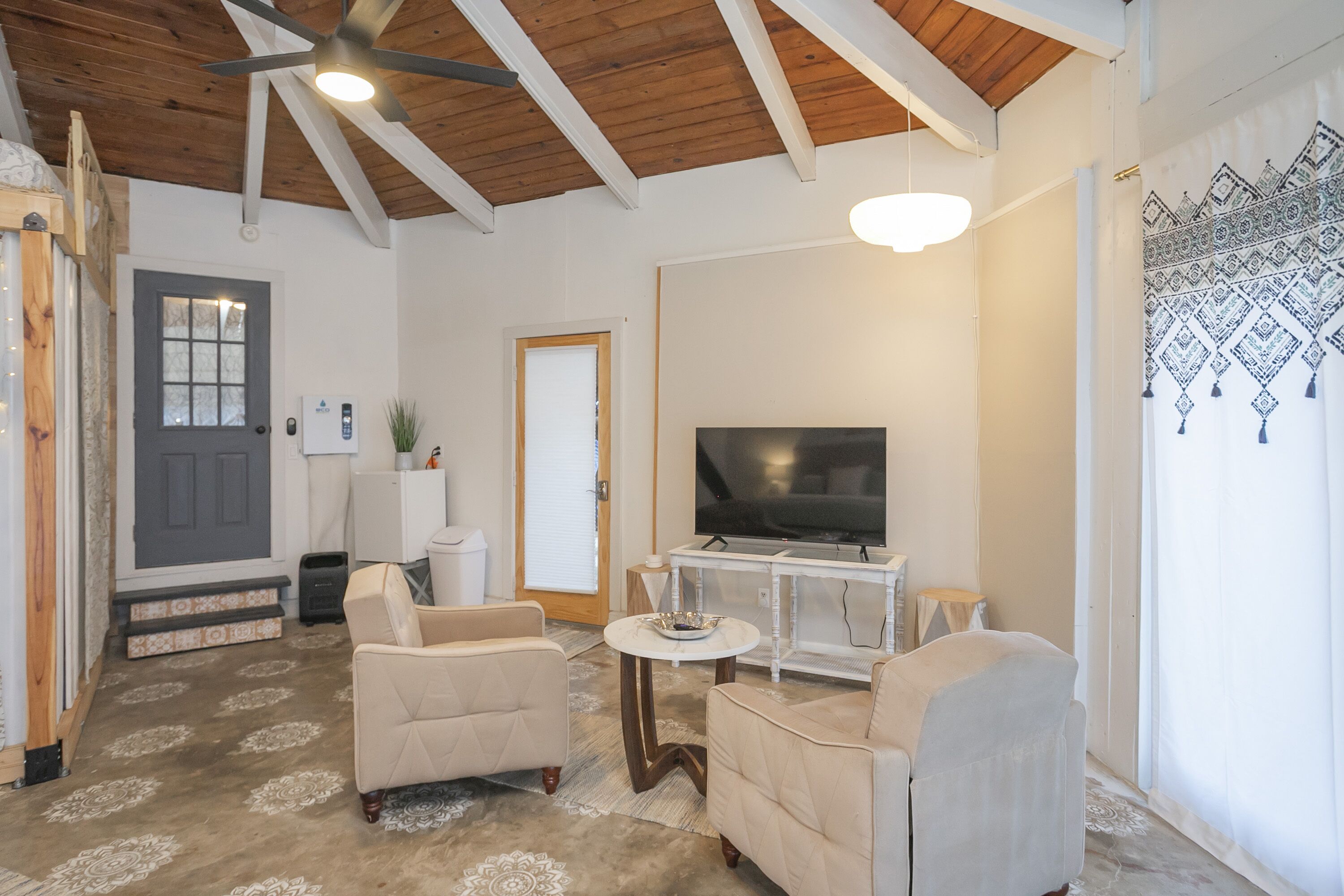 Un salón espacioso con un televisor grande, dos sillones color beige, una mesa de centro y un techo de madera.