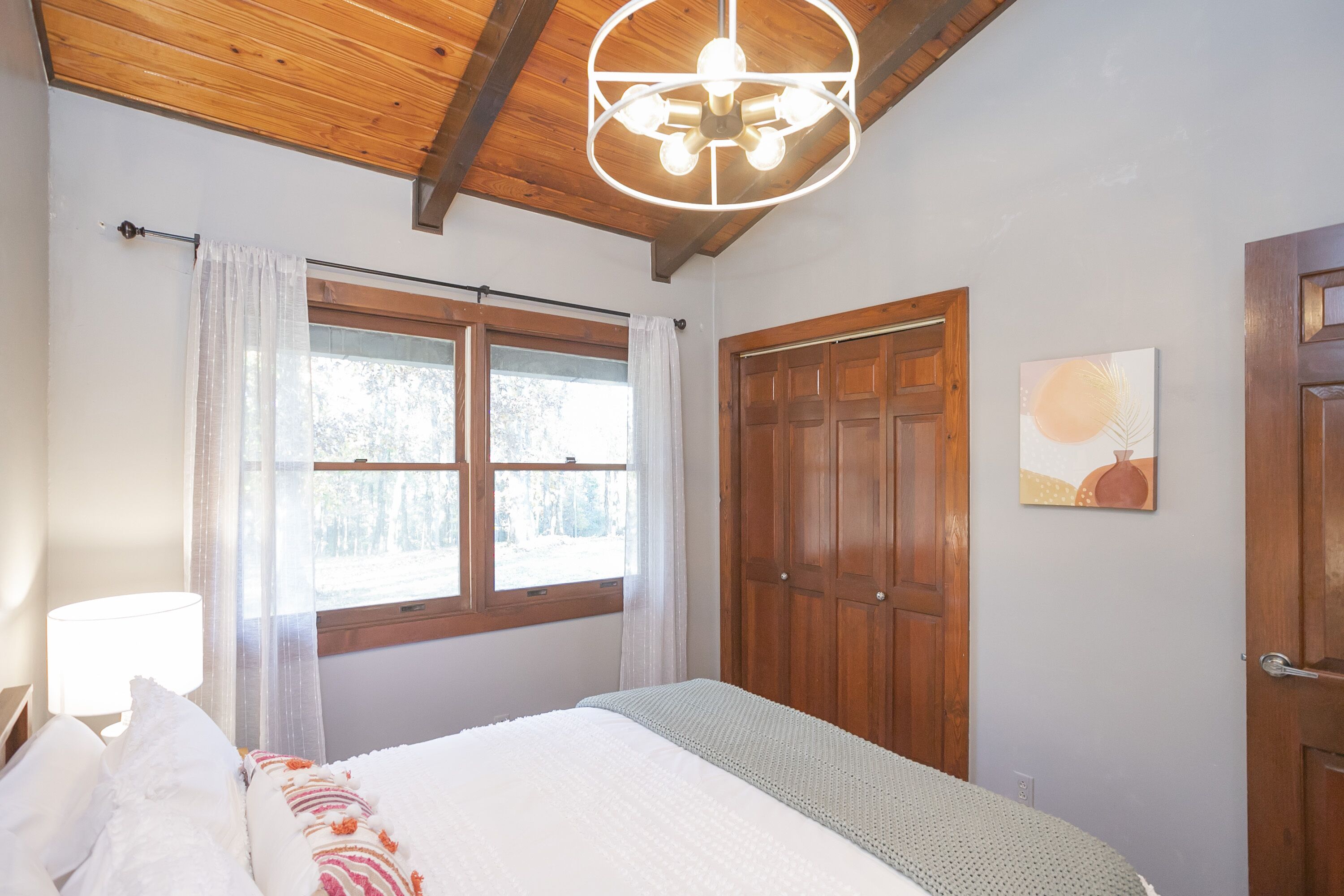 Quarto com teto de madeira, cama com roupa de cama branca, janela com cortinas e porta de madeira.
