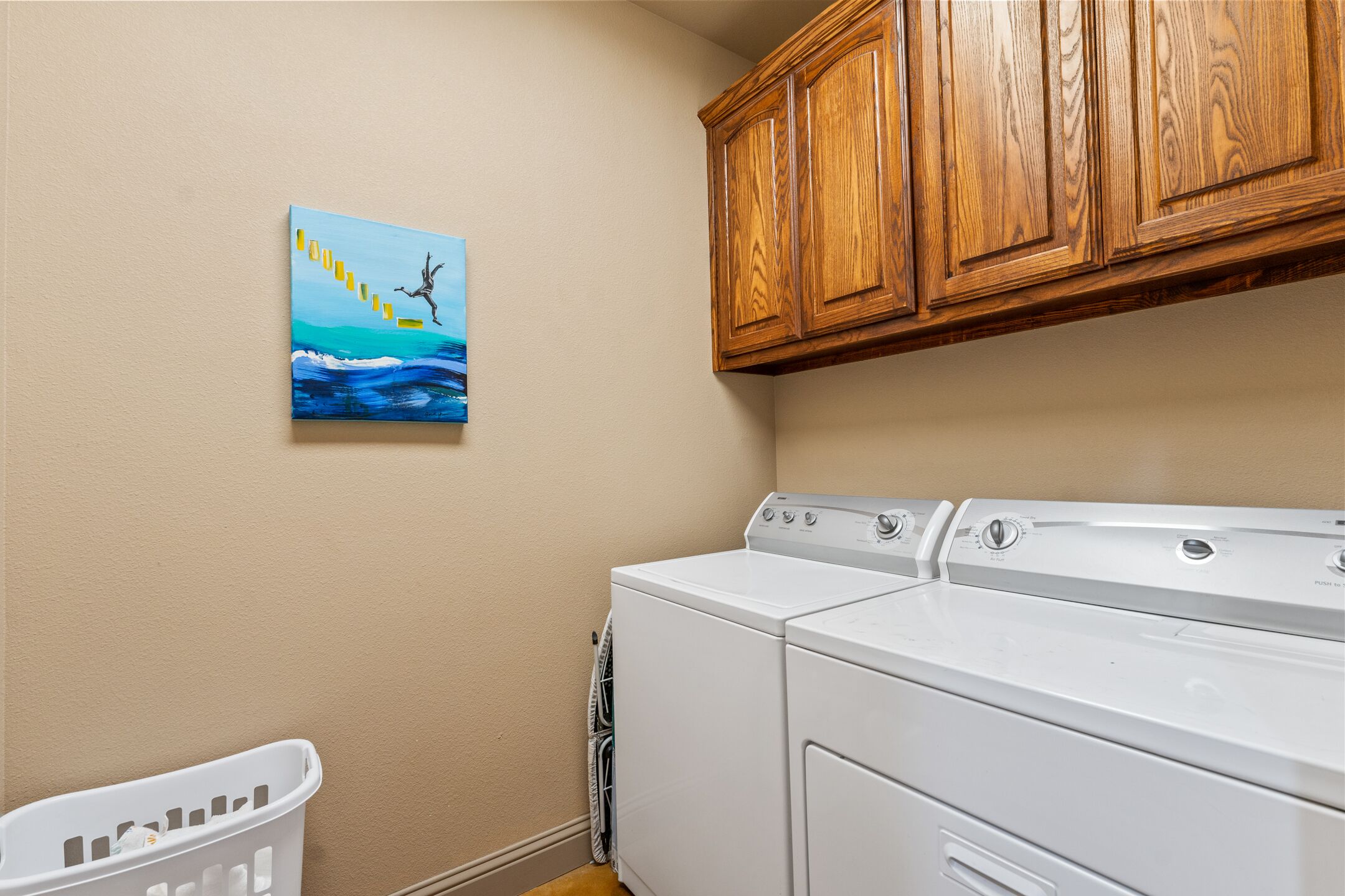 A laundry room with a washing machine, a dryer, wooden cabinets, and a painting on the wall.