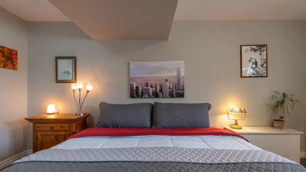 A neatly made bed with a quilted grey blanket, a red throw, and two pillows. There are two bedside tables with lamps, a framed cityscape picture on the wall, and a framed artwork above the bed.