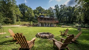 A cabin with a stone fire pit surrounded by Adirondack chairs, set in a lush green landscape.