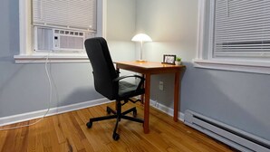 Um quarto com piso de madeira, uma cadeira de escritório preta, uma escrivaninha de madeira, uma luminária e uma pequena planta.