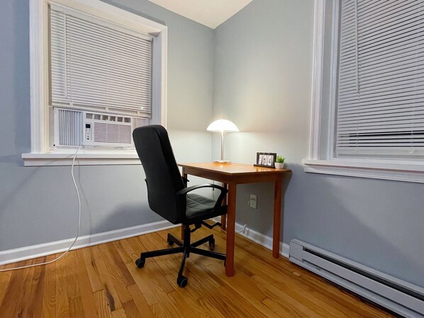 A room with a wooden floor, a black office chair, a wooden desk, a lamp, and a small plant. - Entire place with private parking (Wilmington)