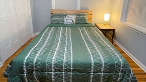 Quarto com uma cama, mesa de cabeceira, janela com persianas e porta.