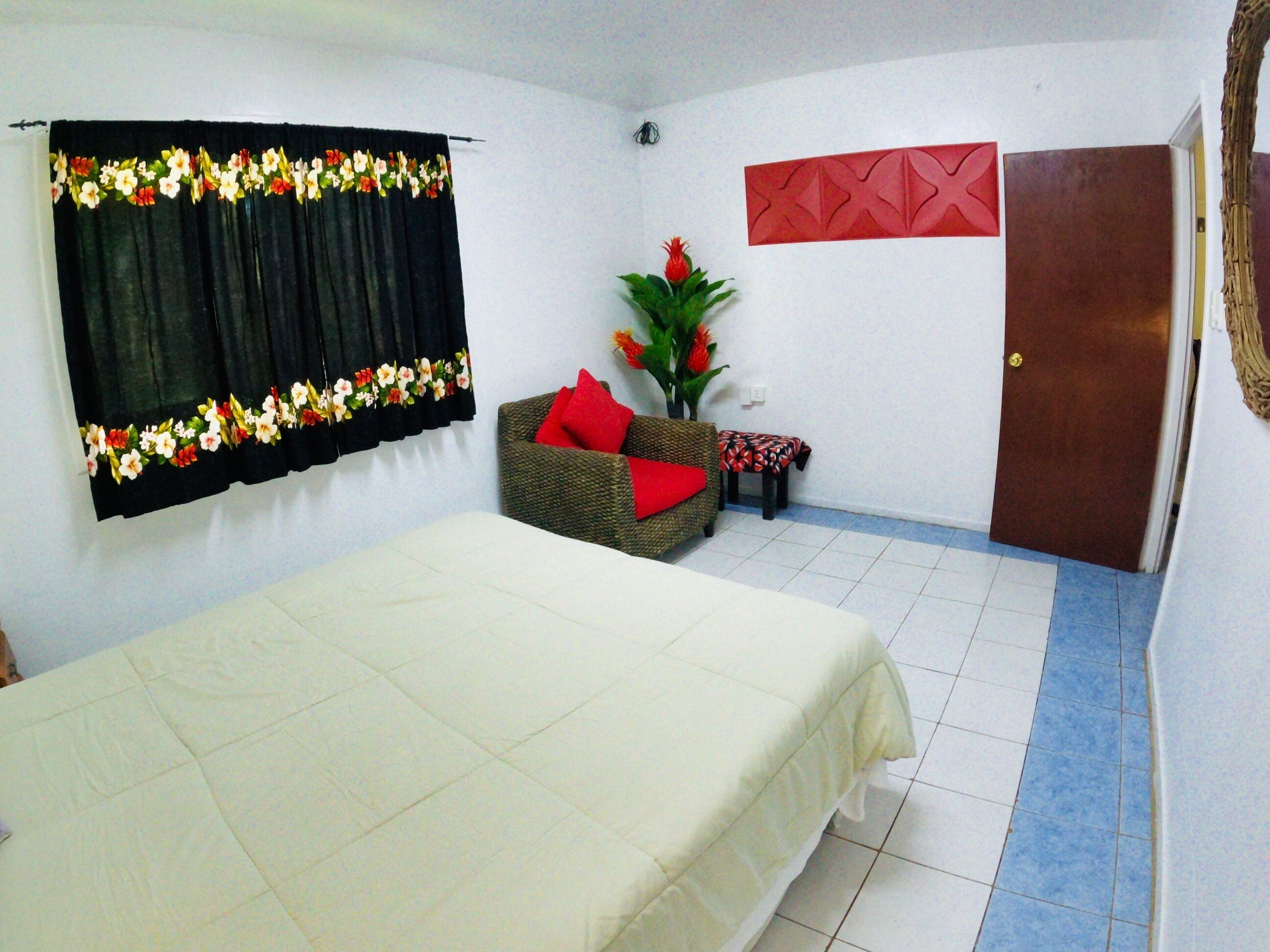 A room with a bed, a wicker sofa with a red cushion, a small table, a potted plant, and a door.