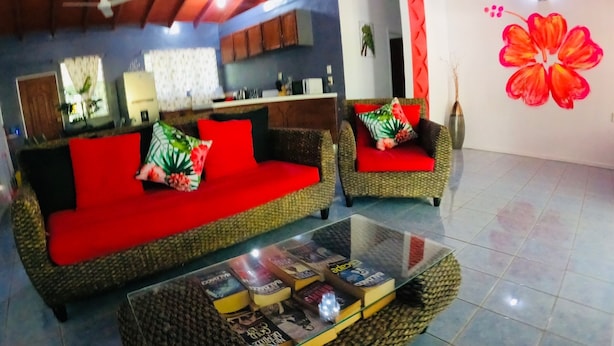A living room with a red sofa and armchair, a glass coffee table with magazines, and a large floral wall decoration. - Cheerful Bungalow with pool! (Apia)