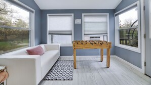 A room with a white sofa, a foosball table, and large windows with blinds.