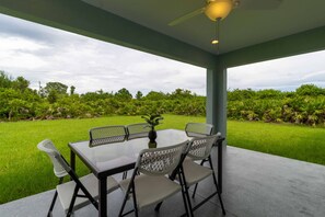 An outdoor patio with a table and chairs, a ceiling fan, and a view of a green lawn and trees.