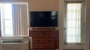 A room with a flat-screen TV on a wooden dresser, a window with blinds, a door with a curtain, and an air conditioning unit. - Historical King George Inn 1 (Niagara-on-the-Lake)