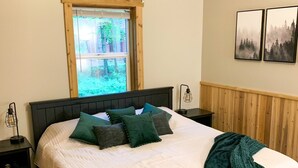 Un dormitorio con una cama grande, un ventanal con vista a un bosque, un ventilador de techo y dos marcos de cuadros colgados en la pared.