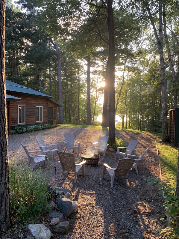 A rustic cabin with a gravel patio, surrounded by tall trees and a fire pit with chairs.