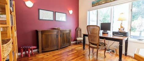 Una habitación con piso de madera, un escritorio con computadora, una silla, una estantería y una ventana con vista a árboles.