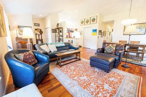 A spacious living room with a blue leather sofa, a matching ottoman, and a wooden coffee table. A patterned rug covers the floor, and the room has hardwood flooring.