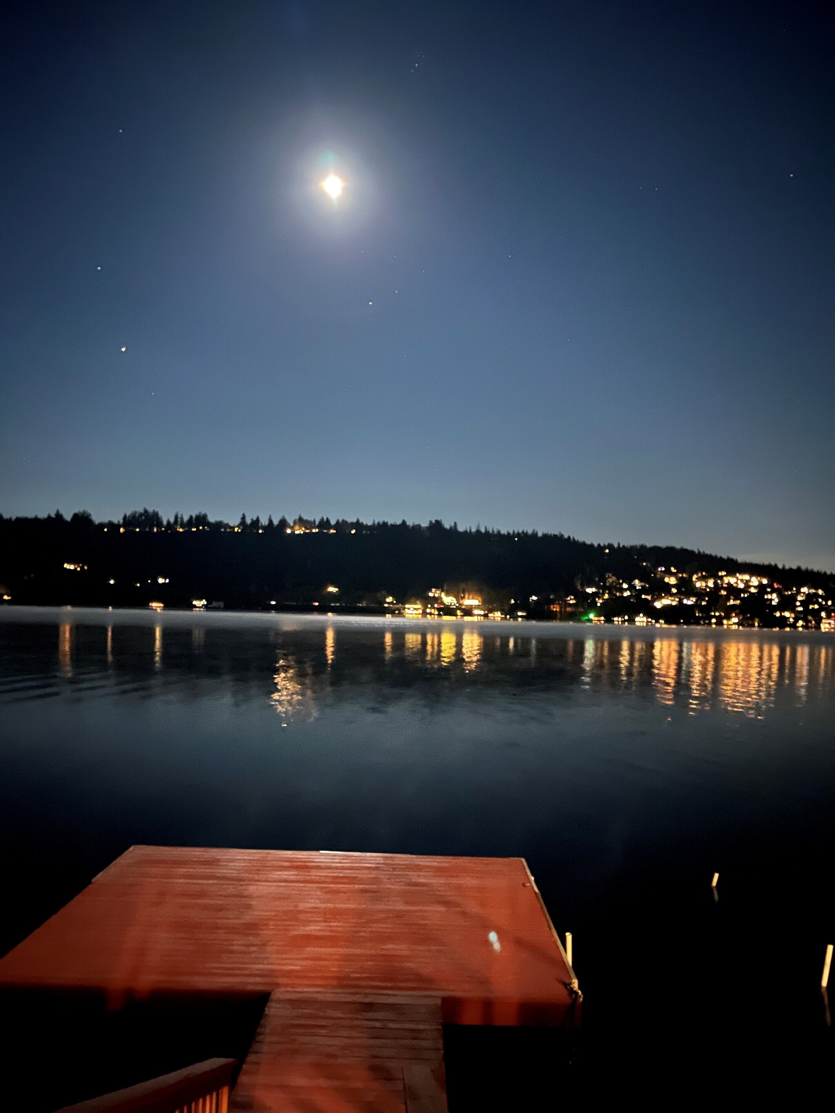 A nighttime view of a calm lake with a wooden dock in the foreground and a cityscape in the background.