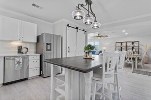 A modern kitchen with white cabinetry, a stainless steel refrigerator, a wooden dining table with white chairs, and a ceiling fan.