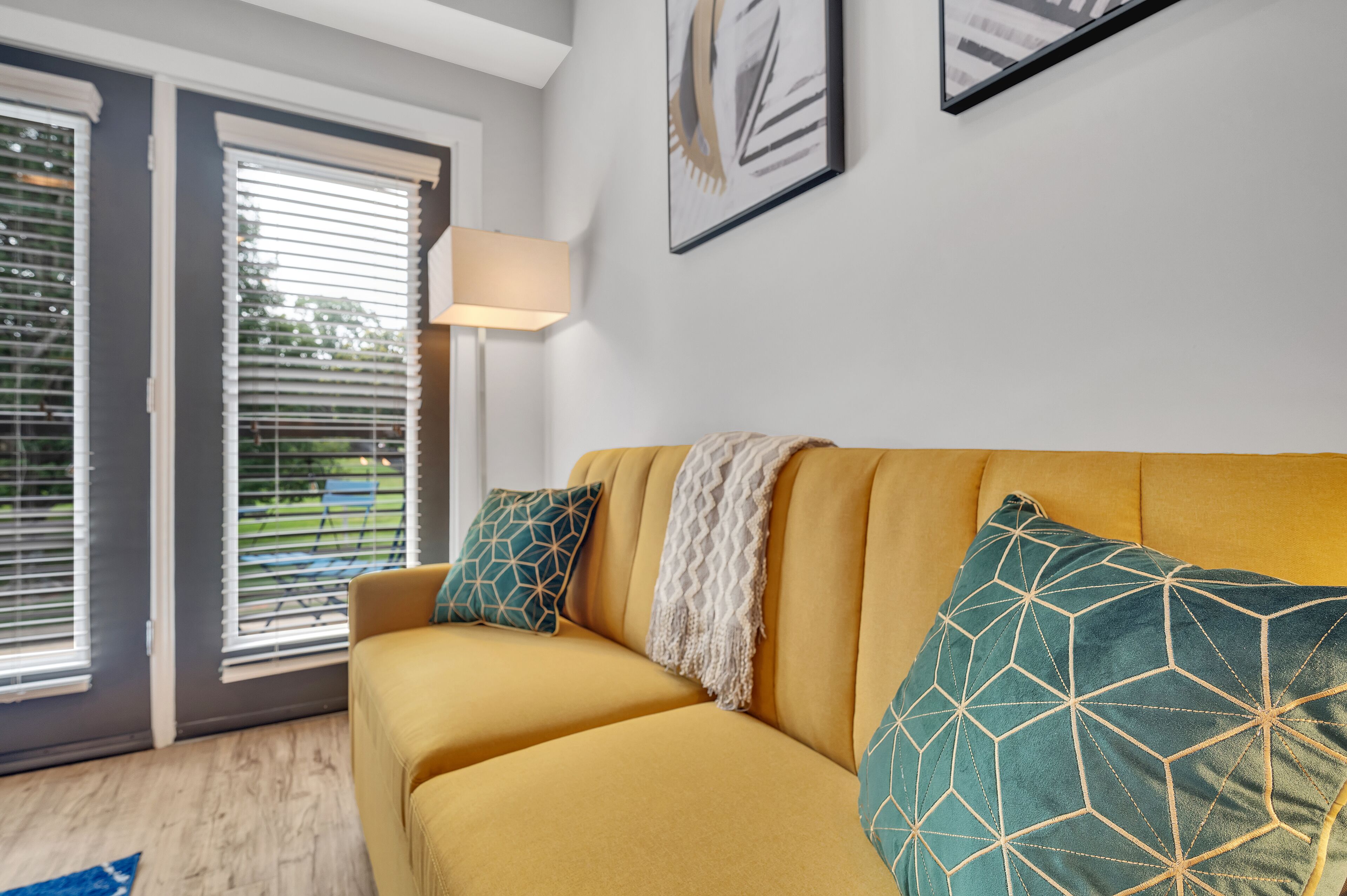 Ein gelbes Sofa mit gemusterten Kissen, einer weißen Decke und einer Stehlampe in einem Zimmer mit einer Schiebetür aus Glas und einem gerahmten Bild an der Wand.