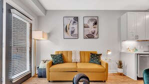 A modern living room with a mustard sofa, abstract wall art, a floor lamp, and a kitchen area with white cabinets. - Modern Luxury ☞ King Bed ☞ 1BR in Plaza Midwood (Charlotte)