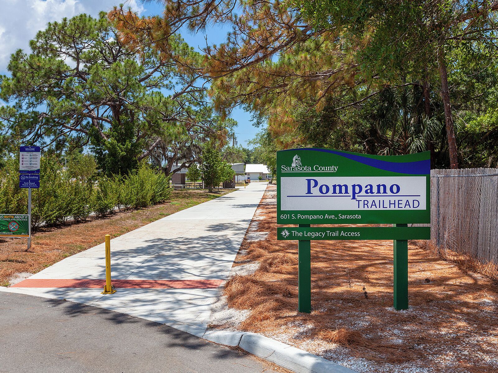 A trailhead sign for Pompano Trailhead in Sarasota County, indicating a welcome area with no smoking and a no parking beach sign.