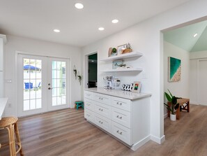 A spacious kitchen with white cabinetry, a coffee station, and a dining area with a wicker chair.