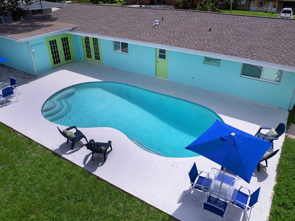 Ein Pool mit gewölbter Kante, umgeben von einer weißen Liegefläche. Blaue Liegestühle und ein blauer Sonnenschirm sind vorhanden, und im Hintergrund ein Gebäude mit grüner Tür und Fenstern.