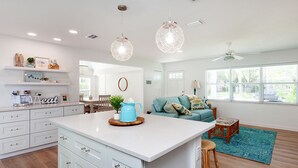 A modern kitchen with white cabinets, a central island, pendant lights, and a dining area with a blue sofa and a wooden coffee table. - Designer Coastal Pool + Game Room home close to Siesta and downtown (Sarasota)