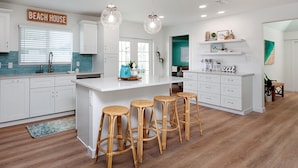 A modern kitchen with white cabinets, a large island with bar stools, and pendant lighting. - Designer Coastal Pool + Game Room home close to Siesta and downtown (Sarasota)