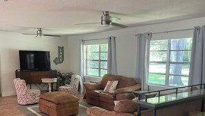 A spacious living room with a large brown sofa, a small round table, a TV, and a ceiling fan. - Canal front pool home to Dora Chain of Lakes (Tavares)