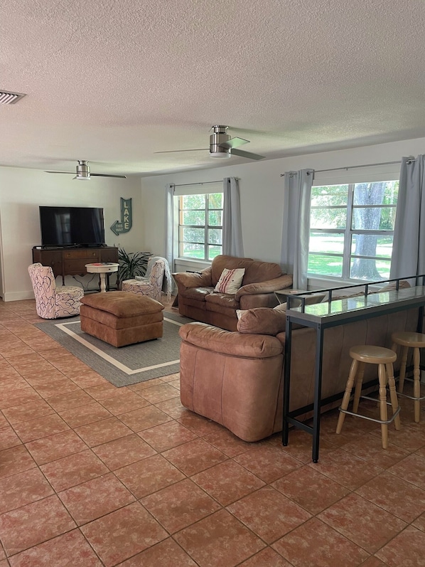 A spacious living room with a large brown sofa, a small round table, a TV, and a ceiling fan. - Canal front pool home to Dora Chain of Lakes (Tavares)