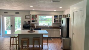 A modern kitchen with white cabinets, a central island with stools, a refrigerator, and a window providing a view of greenery. - Canal front pool home to Dora Chain of Lakes (Tavares)