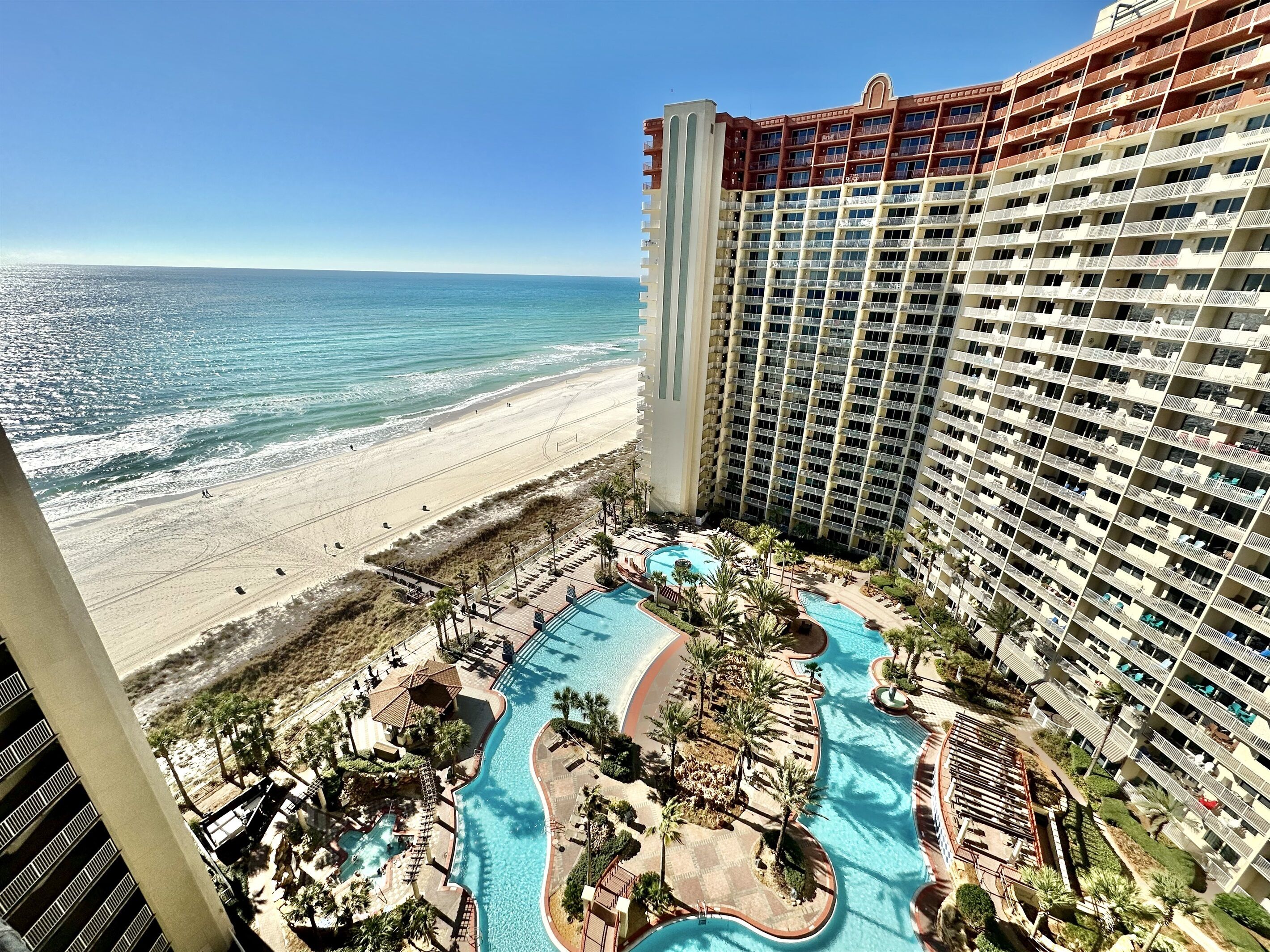 Ein Hochhaus-Hotel mit Blick auf den Strand, ein Poolbereich mit verschiedenen Abschnitten und ein Strand im Hintergrund.