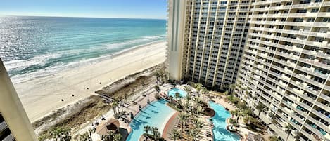 Um hotel alto com vista para a praia, área de piscina com várias seções e uma praia ao fundo.