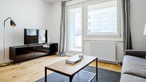 A modern living room with a flat-screen TV on a dark wooden TV stand, a small table with a book, a grey sofa, and a large window with curtains. - Lovely Leopoldstadt 2BR w/ Elevator & city view, nr Danube, by Blueground (Vienna)