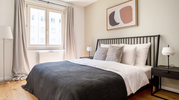A bedroom with a bed, a bedside table, a lamp, a window with curtains, and a wall art piece. - Lovely Leopoldstadt 2BR w/ Elevator & city view, nr Danube, by Blueground (Vienna)
