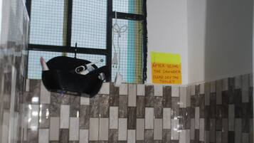 A bathroom with a shower, a window with a mesh screen, a tiled wall, a sign instructing to dry the toilet after using the shower, and a toilet paper roll.