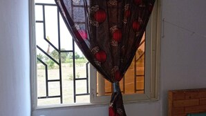 Una habitación con ventana, cabecero de madera y una pequeña mesa con un reloj y un objeto rojo encima.