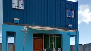 Un edificio de dos pisos con fachada azul, una puerta de madera y un pequeño porche con una banca y una mesa.