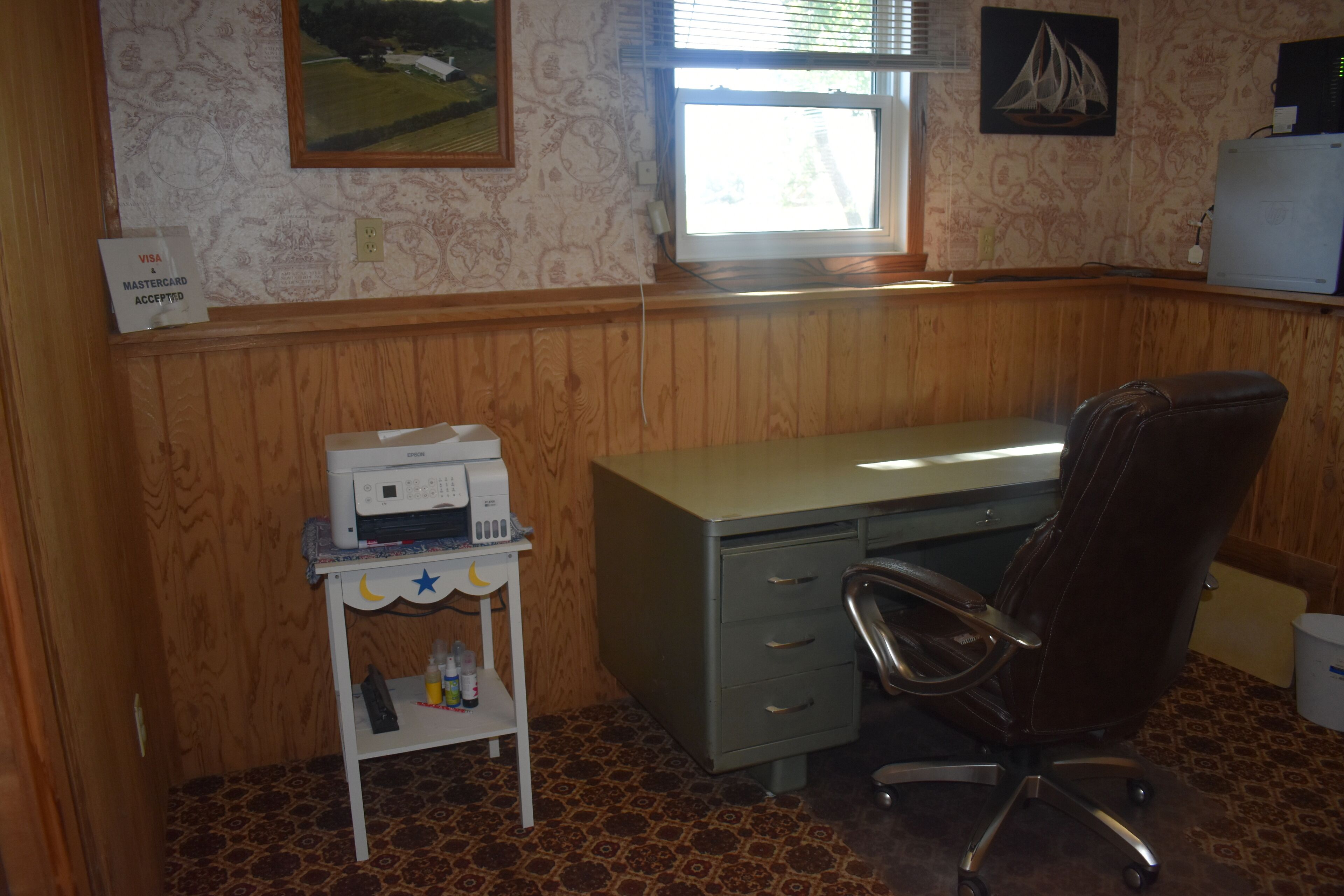 A room with a wooden paneled wall, a desk with a chair, a printer on a small table, and a window with blinds.
