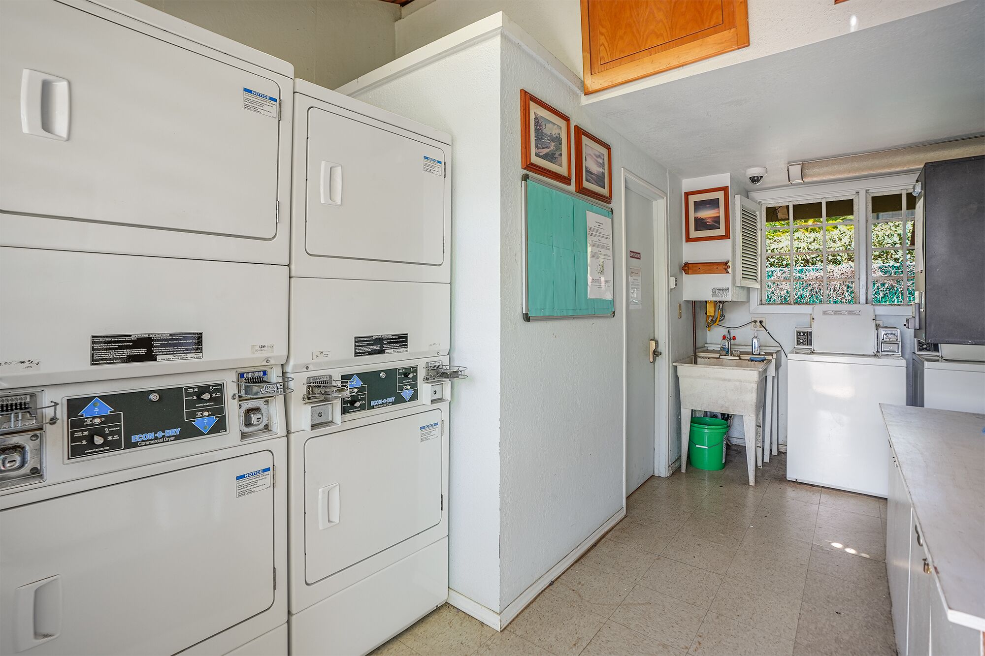 A laundry room with stacked washers and dryers, a small sink, and a window with a view of greenery.