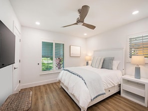 A bedroom with a bed, a ceiling fan, a window with blinds, a bedside table with a lamp, and a TV on the wall.