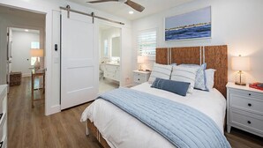 Ein Schlafzimmer mit einem Bett, Nachttischen, einem Deckenventilator, einem Gemälde und einer Tür zu einem Badezimmer.
