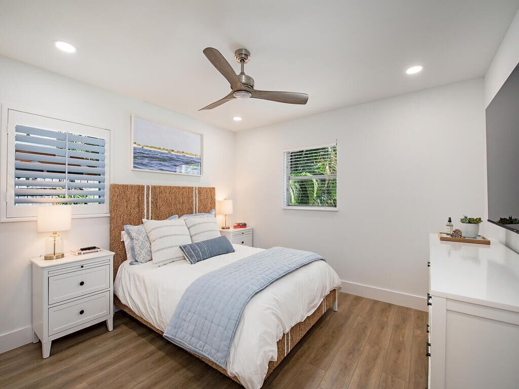 Quarto com cama, mesas de cabeceira, ventoinha no teto, janela com persianas e TV fixada na parede.