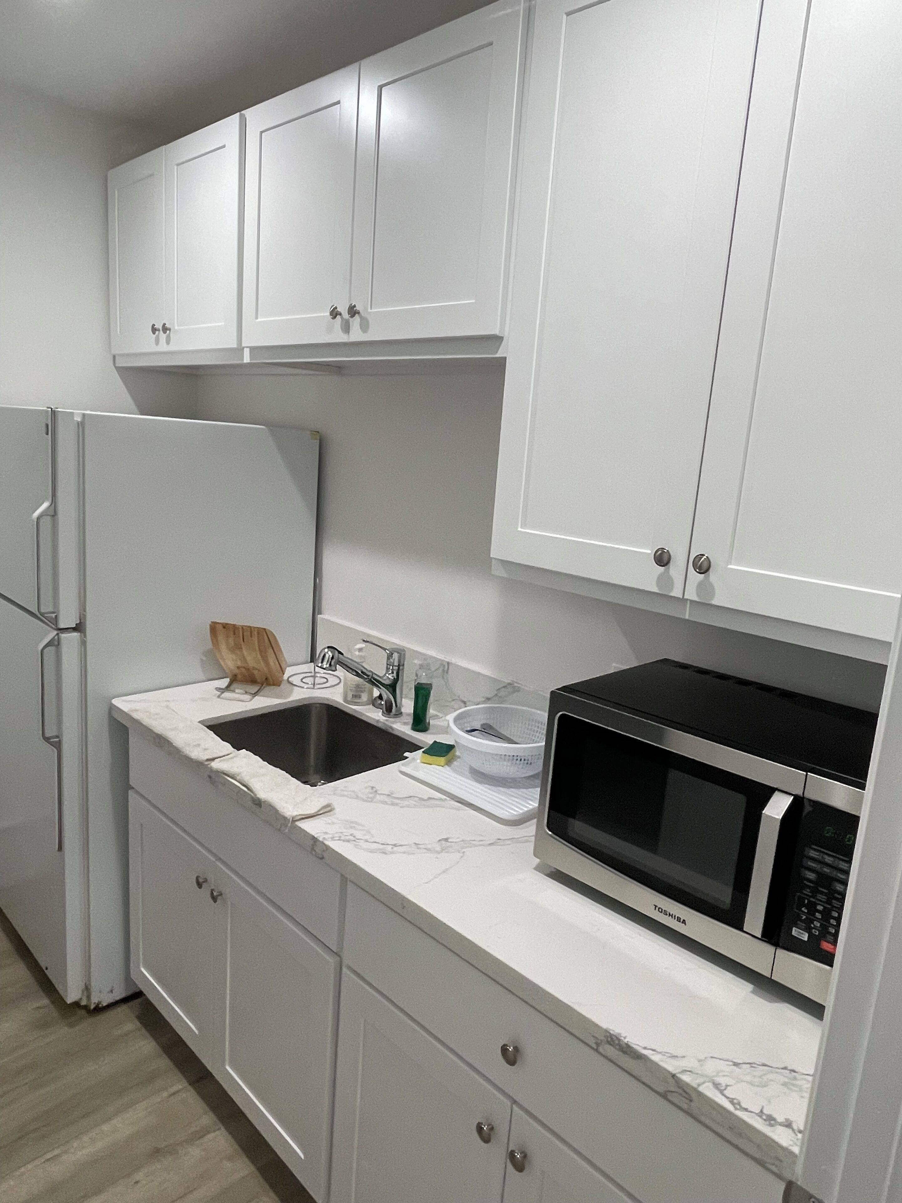 A compact kitchen with white cabinets, a stainless steel refrigerator, a sink, and a microwave.