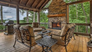 A covered outdoor deck with stone fireplace, wooden furniture, and a grill. - Songbird Ridge - Aska Adventure Area | Panoramic Views | Hot Tub | Outdoor Fireplace (Blue Ridge)