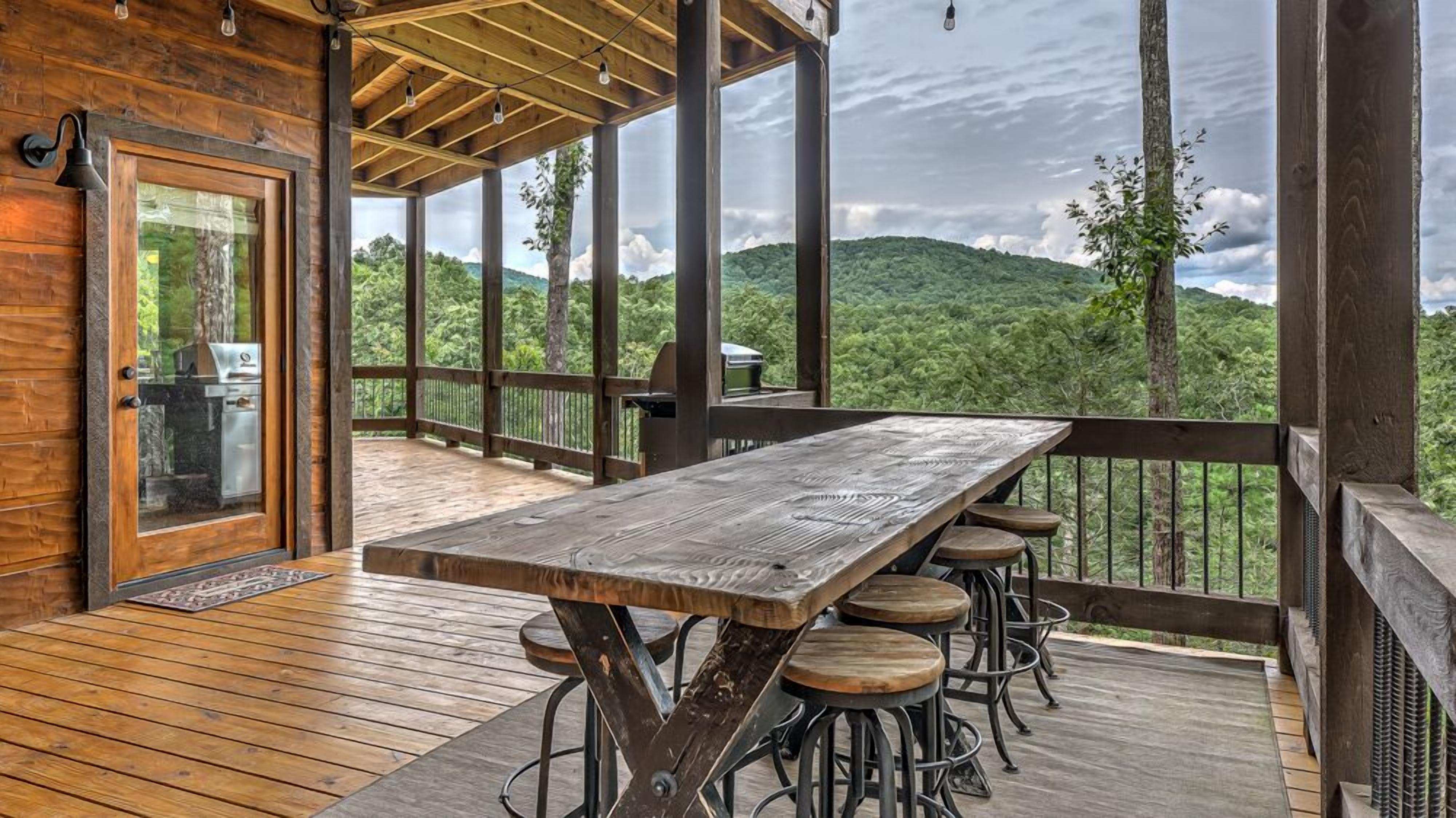A wooden deck with a large table and stools, overlooking a forested landscape.